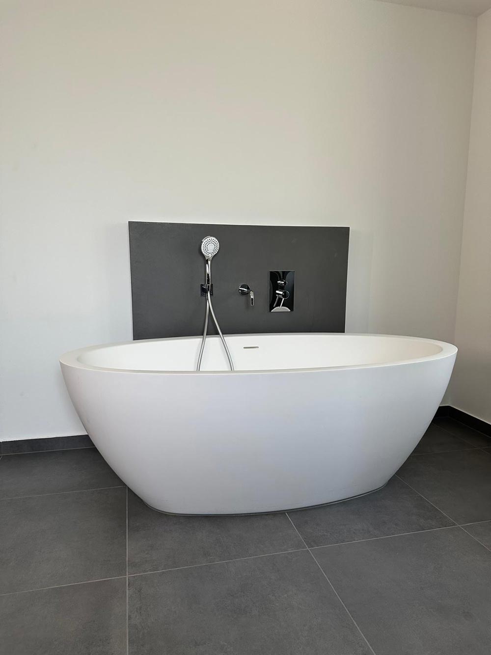 Moderne weiße Badewanne vor grauer Wand mit Armatur und Fliesenboden