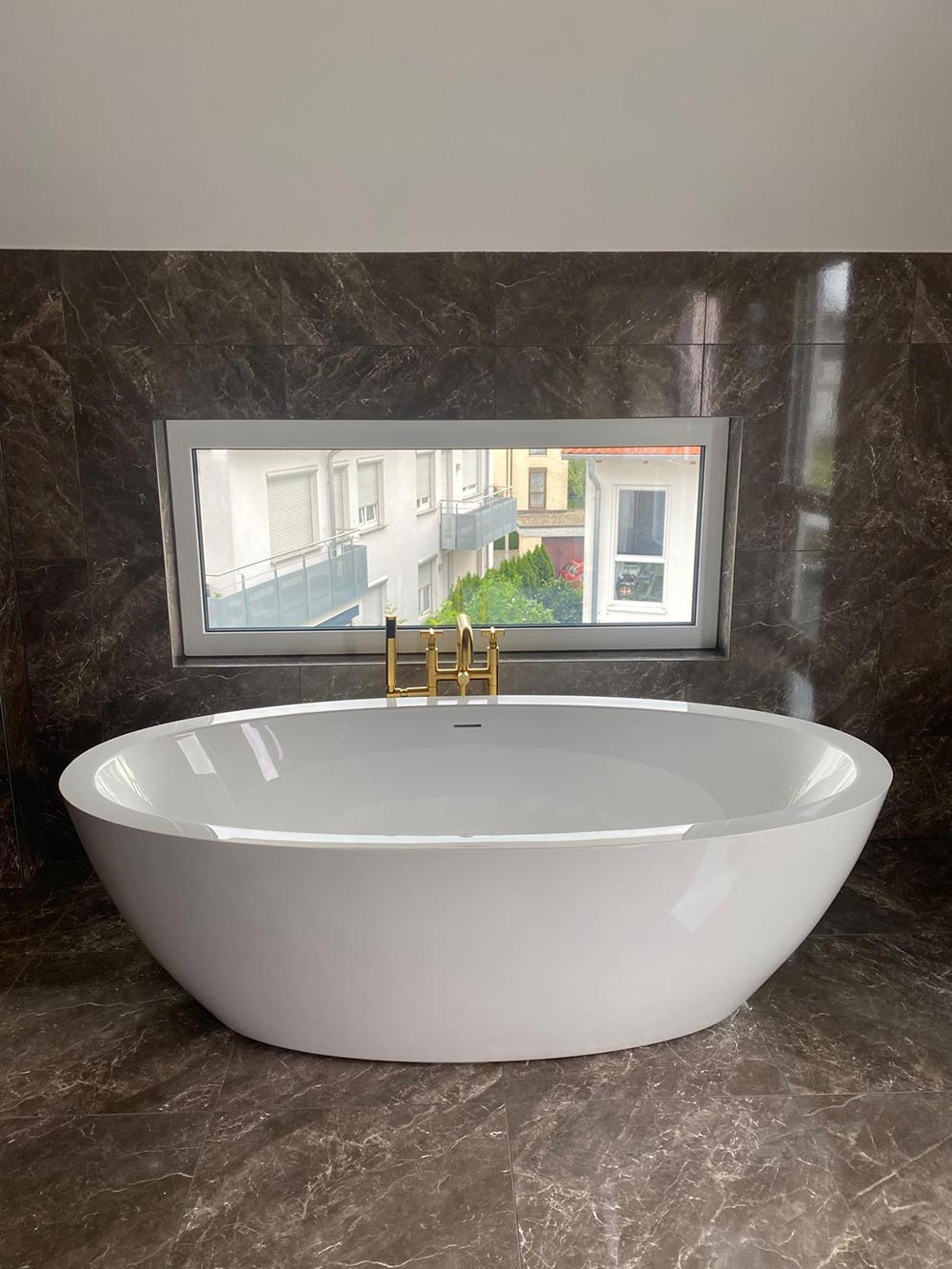 Weisse ovale Badewanne vor dunkler Marmorwand mit Fenster und Goldhahn
